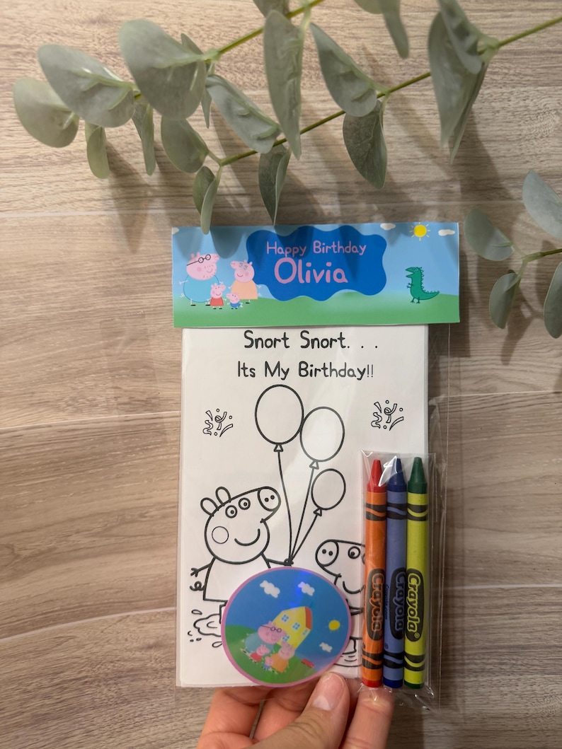 Peut inclure: Un ensemble de coloriage sur le th&egrave;me de l'anniversaire avec des personnages de Peppa Pig. L'ensemble comprend un livre de coloriage avec le texte "Snort Snort... It's My Birthday!!" et "Happy Birthday Olivia", des crayons et un autocollant rond. Les crayons sont orange, bleu et vert.