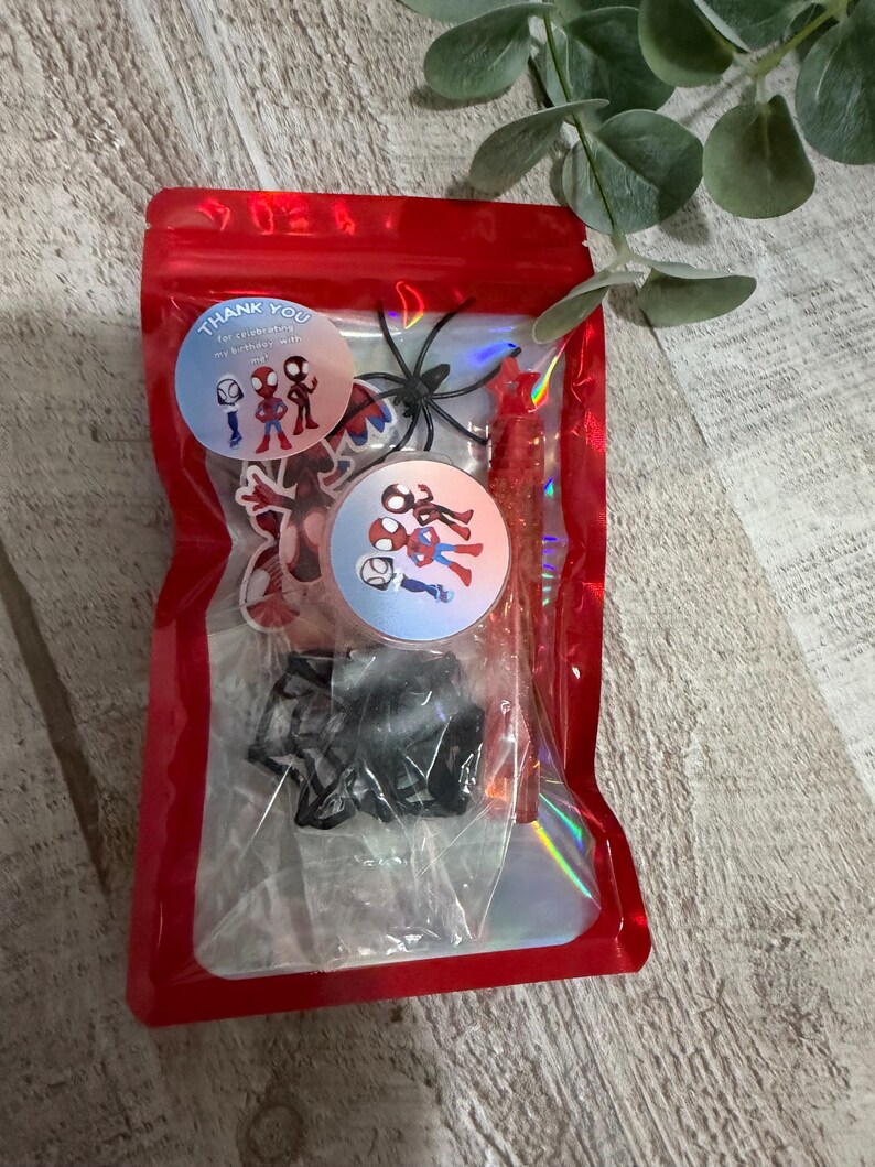 May include: A red cellophane bag filled with party favors. Inside are stickers with Spider-Man characters, a black plastic spider, and a red candy stick. The bag has a holographic sheen and the text "Thank You".