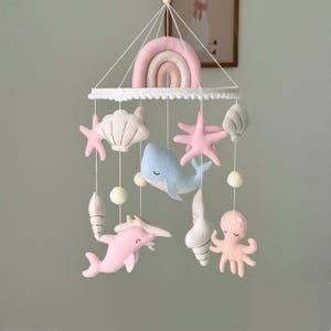 Peut inclure: Un mobile suspendu composé d'animaux marins en feutrine et d'un arc-en-ciel. Le mobile comprend une baleine bleu clair, un dauphin rose, une pieuvre rose, des étoiles de mer et divers coquillages suspendus par des fils blancs à un cadre circulaire.