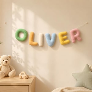 May include: A decorative wall hanging featuring plush, stuffed fabric letters spelling the name OLIVER in various pastel colours including green, orange, cream, blue, yellow, and pink, suspended on a thin white string against a plain wall.
