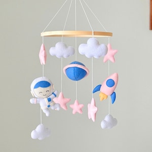 Peut inclure: Mobile pour bébé fait à la main avec un astronaute, une fusée, des étoiles, des nuages et une planète. L'astronaute est blanc avec des détails bleus, la fusée est rose avec des détails bleus et jaunes. Suspendu à un cercle en bois.