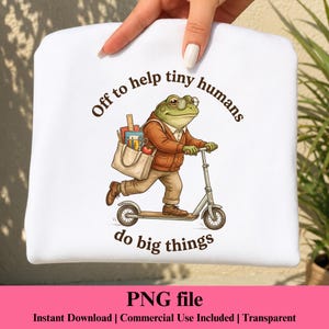 Puede incluir: Una camiseta blanca con el gráfico de una rana verde con gafas y cárdigan marrón, montada en un patinete y llevando una bolsa con material escolar. El texto dice, Off to help tiny humans do big things.