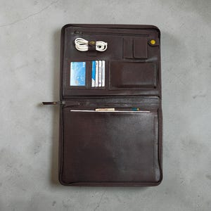 Könnte beinhalten: Geöffnete braune Ledermappe mit mehreren Fächern. Merkmale sind ein Reißverschluss, Kartenfächer und ein Stifthalter. Enthält ein weißes Ladekabel und einen gelben Knopfverschluss.