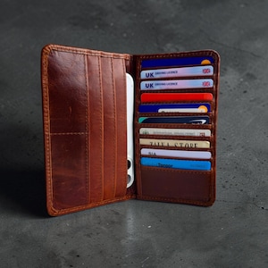 May include: Open brown leather wallet with multiple card slots filled with cards and a smartphone. The wallet is open, revealing its interior and the items stored within. The leather has a textured appearance.