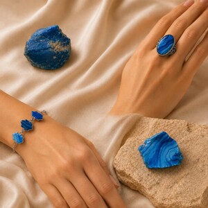 Op de afbeelding: Een zilveren ring met een ovale blauwe steen met banden en een zilveren kettingarmband met drie kleine blauwe steenbedels. Twee ruwe, grillige blauwe agaatstenen zijn aanwezig, één rustend op een getextureerde beige steen en één op stof.