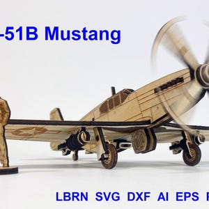 May include: A laser-cut wooden model of a P-51B Mustang airplane with a spinning propeller and a standing pilot figure. The text P-51B Mustang appears in blue at the top, and LBRN SVG DXF AI EPS PDF CDR is printed in blue at the bottom.