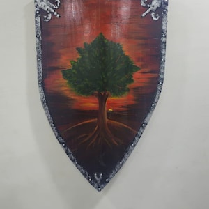 May include: Shield-shaped wall art depicting a tree with a green crown and roots against a sunset. The wooden shield has a dark border with decorative silver accents. The artwork is mounted on a wall.