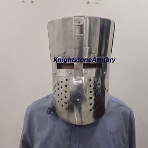 May include: A polished silver knight's helmet with a rectangular eye slit and numerous ventilation holes. The helmet is riveted together and has a flat top. The text "KnightstoneArmory" is visible on the helmet.