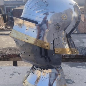 Könnte beinhalten: Ein polierter Ritterhelm aus Stahl mit dekorativen goldfarbenen Verzierungen. Der Helm hat ein Visier, einen Nackenschutz und verzierte florale Designs. Das Metall reflektiert die Umgebung und zeigt die Handwerkskunst.