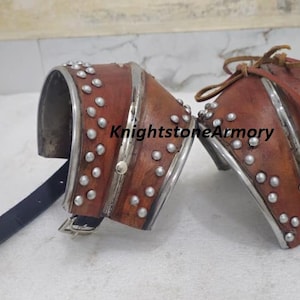 Puede incluir: Par de brazales de cuero marrón con detalles y remaches de metal plateado. Cada protector tiene una forma curva y una correa. El texto "KnightstoneArmory" es visible en el brazalete derecho.