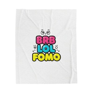 May include: White blanket with the text "BRB LOL FOMO" in colorful bubble letters. The text is arranged vertically with a cartoon face above the text. The blanket has a slightly wrinkled texture.