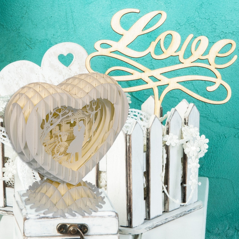 Peut inclure: Un c&oelig;ur en papier blanc avec un design 3D pop-up d'un couple debout devant un gazebo. Le c&oelig;ur est pos&eacute; sur une bo&icirc;te blanche avec un couvercle en bois. Le mot "Love" est &eacute;crit en cursive sur une pancarte en bois derri&egrave;re le c&oelig;ur.