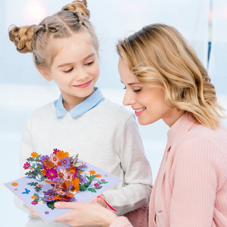 Peut inclure: Une jeune fille offre une carte pop-up 3D avec un bouquet de fleurs color&eacute;es &agrave; une femme. La carte est ouverte et les fleurs sont en pleine floraison. La carte est de couleur violet clair avec des feuilles vertes et un papillon.