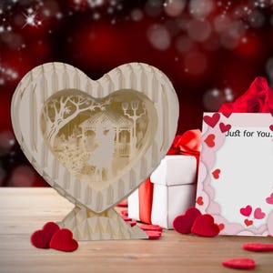 Peut inclure: Une bo&icirc;te en papier en forme de c&oelig;ur blanche avec une sc&egrave;ne pop-up 3D d'un couple se tenant dans un parc. La bo&icirc;te est pos&eacute;e sur une table en bois avec un ruban rouge et une bo&icirc;te cadeau blanche &agrave; c&ocirc;t&eacute;. Une carte avec le texte "Just for You." est &eacute;galement sur la table.