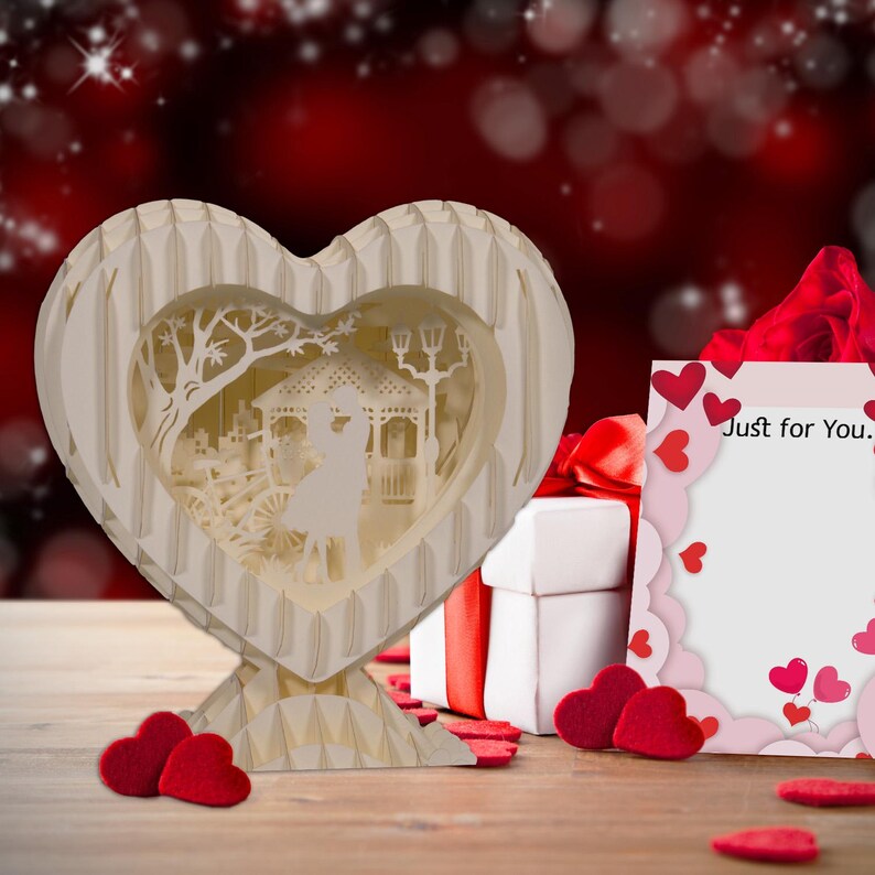 Peut inclure: Une bo&icirc;te en papier en forme de c&oelig;ur blanche avec une sc&egrave;ne pop-up 3D d'un couple se tenant dans un parc. La bo&icirc;te est pos&eacute;e sur une table en bois avec un ruban rouge et une bo&icirc;te cadeau blanche &agrave; c&ocirc;t&eacute;. Une carte avec le texte "Just for You." est &eacute;galement sur la table.