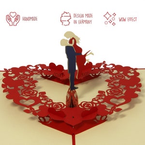 May include: A red and white 3D pop-up card depicting a couple embracing. The card is decorated with a heart-shaped wreath of roses and hearts. The card has the text "Handmade" and "Design made in Germany" on the front.