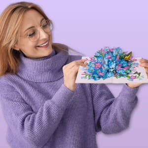May include: A woman is holding a 3D pop-up card with a bouquet of blue forget-me-not flowers and a yellow butterfly.