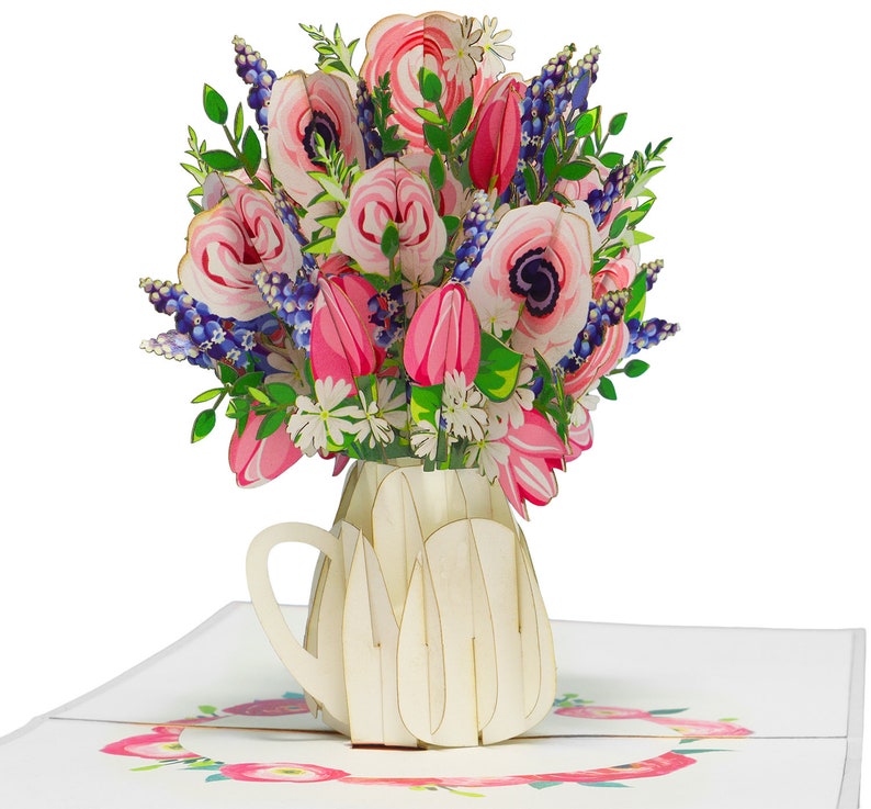 May include: A 3D pop-up greeting card featuring a bouquet of pink tulips, anemones, and blue flowers in a cream-colored vase. The card is open, revealing a floral wreath design. The card is made of paper.