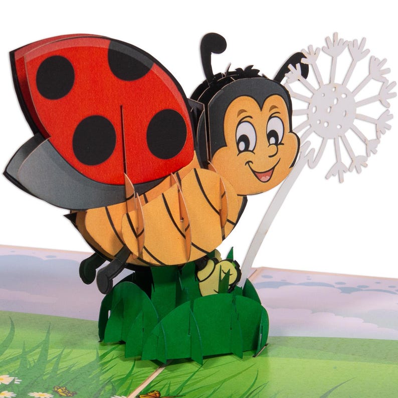 May include: A 3D pop-up card featuring a ladybug with a red and black shell, holding a white dandelion. The ladybug is smiling and has a yellow and orange body. The card is set against a green grassy background.