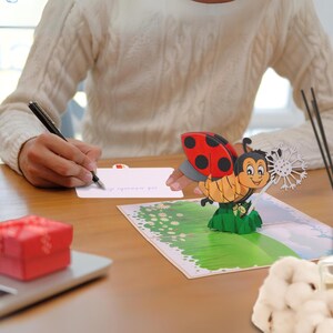 May include: A 3D pop-up card featuring a ladybug with red and black wings, standing on a green leaf with a dandelion in the background. The card is open and a person is writing on the inside.
