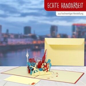 May include: A 3D pop-up card featuring a cityscape with a red church, a blue ship, and a red lion. The card is open and the cityscape is raised. The card is on a wooden surface. The card is accompanied by a yellow envelope. The text "Echte Handarbeit" is on the envelope, which translates to "Real Handcraft". The text "aus hochwertiger Herstellung" is also on the envelope, which translates to "from high-quality production".