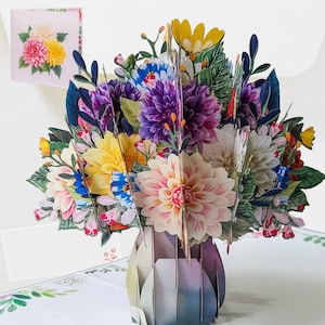 Può includere: Un biglietto pop-up 3D con un vivace bouquet di fiori di carta in un vaso. I fiori includono fiori rosa, gialli, viola e blu con foglie verdi. È visibile un biglietto più piccolo con un design floreale simile.
