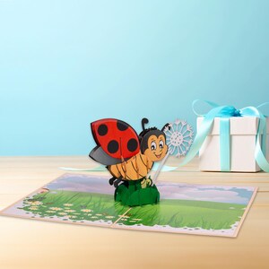 May include: A 3D pop-up card featuring a ladybug with red and black spots and a yellow and black body. The ladybug is standing on a green leaf with a white dandelion in the background.