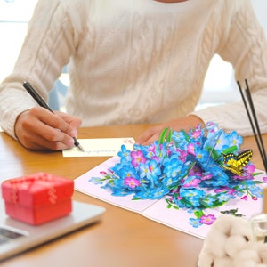 May include: A 3D pop-up card featuring a bouquet of blue and pink forget-me-not flowers with a yellow butterfly perched on top. The card is open and the flowers are in full bloom.