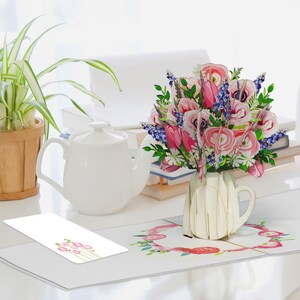 May include: A 3D paper bouquet in a white vase, featuring pink tulips, roses, and blue flowers. A white teapot and a potted plant are on the desk. A card with pink flowers is on the left.