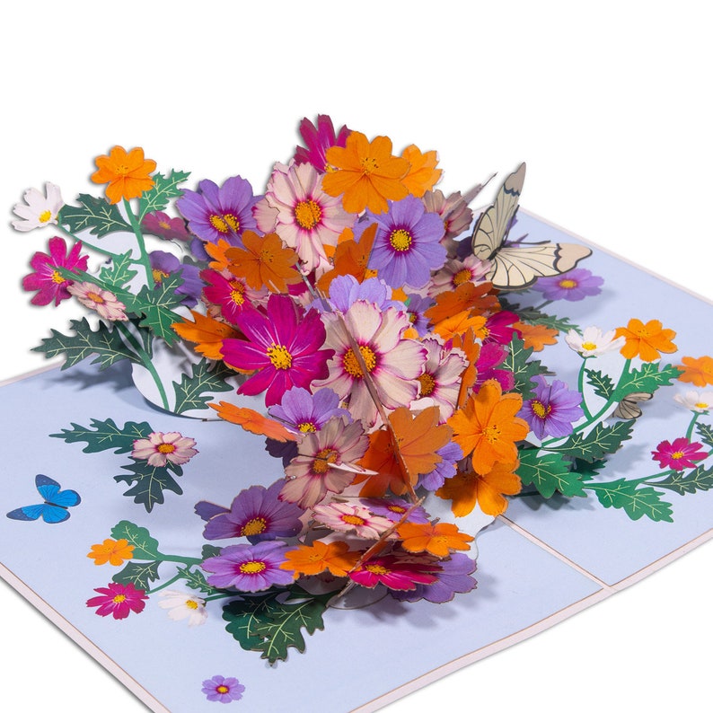 Peut inclure: Une carte pop-up 3D repr&eacute;sentant un bouquet de fleurs color&eacute;es, dont des roses, des violettes, des oranges et des blanches. Les fleurs sont dispos&eacute;es dans un vase et entour&eacute;es de feuilles vertes. Un papillon vole au-dessus des fleurs.