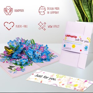 May include: A 3D pop-up card featuring a bouquet of blue forget-me-not flowers with a yellow butterfly. The card is open and the flowers are popping up. The card is on a white surface. The card has a pink envelope with the text "Just for you." on it. The card is handmade and plastic-free. The card is designed and made in Germany.