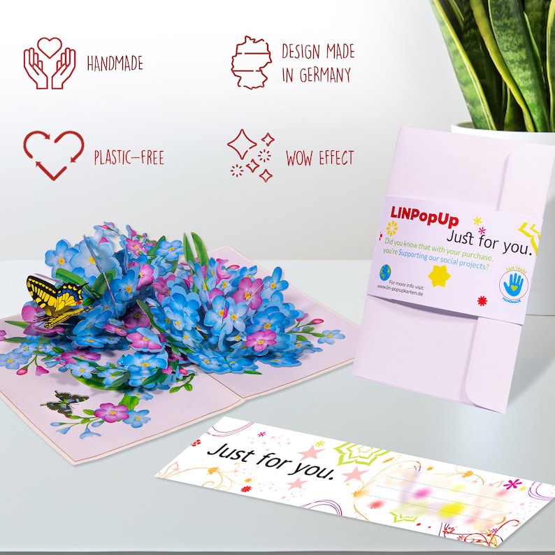 May include: A 3D pop-up card featuring a bouquet of blue forget-me-not flowers with a yellow butterfly. The card is open and the flowers are popping up. The card is on a white surface. The card has a pink envelope with the text "Just for you." on it. The card is handmade and plastic-free. The card is designed and made in Germany.