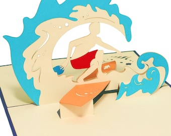 May include: A 3D pop-up card featuring a surfer riding a wave. The surfer is wearing red shorts and is silhouetted against a blue wave. The card is made of paper and has a cream background.