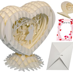 Peut inclure: Une carte pop-up 3D blanche en forme de c&oelig;ur. La carte repr&eacute;sente un couple debout devant un kiosque avec un v&eacute;lo en arri&egrave;re-plan. La carte est livr&eacute;e avec une enveloppe blanche et une carte rose avec les mots "Just for You." imprim&eacute;s dessus.