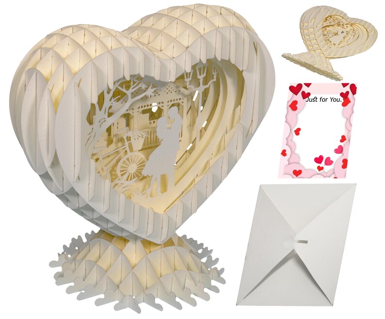 Peut inclure: Une carte pop-up 3D blanche en forme de c&oelig;ur. La carte repr&eacute;sente un couple debout devant un kiosque avec un v&eacute;lo en arri&egrave;re-plan. La carte est livr&eacute;e avec une enveloppe blanche et une carte rose avec les mots "Just for You." imprim&eacute;s dessus.