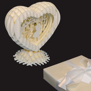 Peut inclure: Une sculpture en papier blanc en forme de c&oelig;ur avec un design d&eacute;taill&eacute; d'un couple se tenant dans un parc. La sculpture est sur une base en papier blanc.
