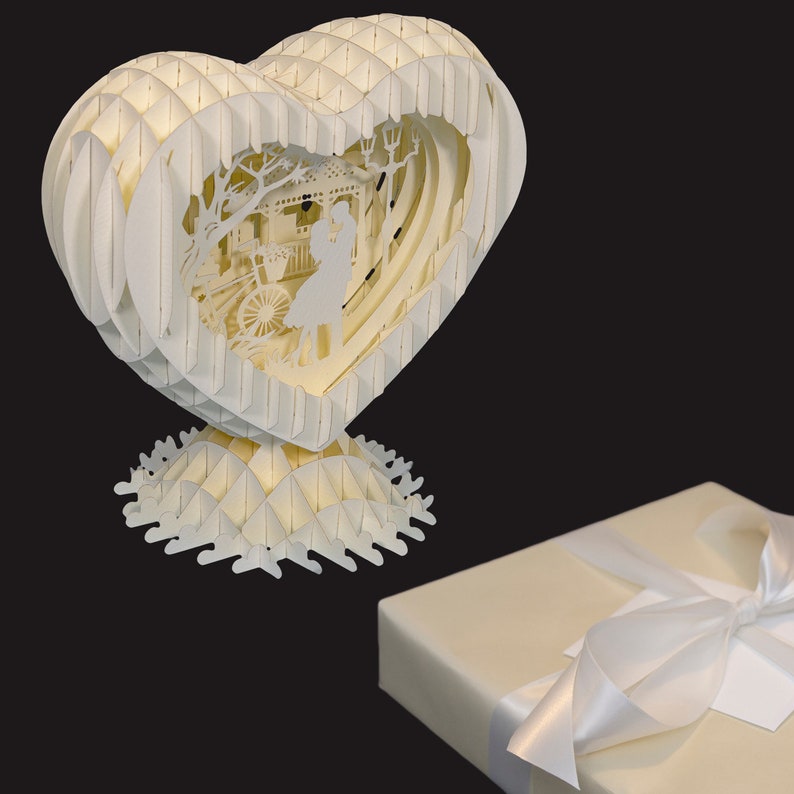 Peut inclure: Une sculpture en papier blanc en forme de c&oelig;ur avec un design d&eacute;taill&eacute; d'un couple se tenant dans un parc. La sculpture est sur une base en papier blanc.
