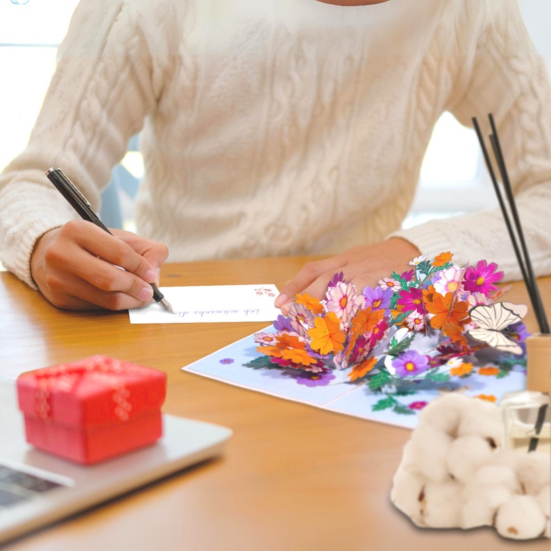 Peut inclure: Une personne &eacute;crit un message sur une carte blanche avec un stylo noir. La carte est &agrave; c&ocirc;t&eacute; d'une carte pop-up 3D avec un bouquet de fleurs color&eacute;es et un papillon. La carte est sur une table en bois.