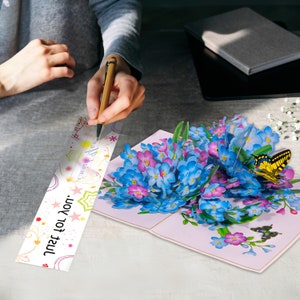 May include: A 3D pop-up card featuring a bouquet of blue and purple forget-me-not flowers with two butterflies, one yellow and one blue. The card has a white insert with the text "Just for you."