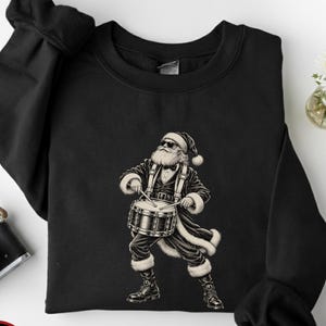 May include: A black crewneck sweatshirt featuring a graphic print of Santa Claus wearing sunglasses and a tuxedo while playing a snare drum. The design is rendered in white and gray tones, showcasing Santa with a beard and hat.
