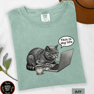 May include: A sage green t-shirt featuring a graphic of a gray cat wearing glasses, sitting at a laptop with a steaming cup of coffee. A speech bubble above the cat contains the text "This is my life." The shirt has a Comfort Colors brand label.