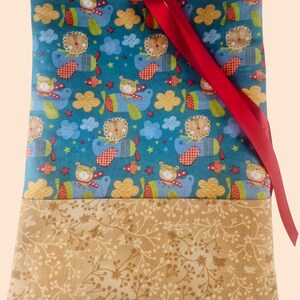 May include: A fabric tote bag with a whimsical design. The top section has a teal background with cartoon bears in aeroplanes, clouds, and stars. The bottom section is tan with a floral pattern. A red ribbon is attached.