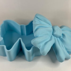 Pode incluir: Uma caixa de bugigangas azul clara em forma de laço, com tampa a condizer. A caixa está aberta, revelando o interior. A tampa é um design de laço detalhado. A caixa é feita de um material sólido e assenta numa superfície branca.