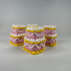Puede incluir: Cuatro bizcochos en miniatura de color rosa, amarillo y blanco. Cada bizcocho tiene tres capas, decoradas con glaseado amarillo en los bordes, detalles de glaseado blanco y glaseado blanco en la parte superior. Los bizcochos están colocados sobre una superficie blanca.