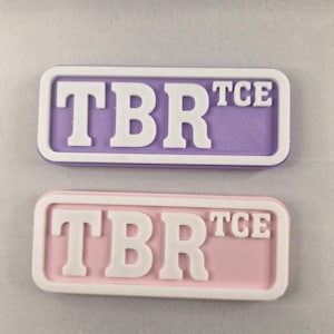 Könnte beinhalten: Zwei rechteckige, pastellfarbene Plaketten mit weißer Schrift. Die obere Plakette ist lavendelfarben mit weißem Text "TBR TCE". Die untere Plakette ist rosa mit weißem Text "TBR TCE". Beide Plaketten haben einen weißen Rand.