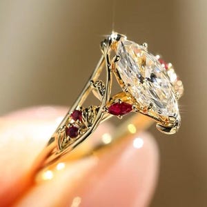 May include: A gold ring featuring a large marquise-cut clear gemstone as the center stone. The band is designed with intricate vine-like metalwork and is accented with several small, deep red marquise and round-cut rubies set along the sides.