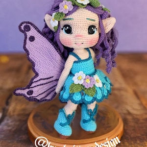 May include: A handcrafted crochet fairy doll with purple hair, a blue dress, and matching boots. The doll features large eyes, rosy cheeks, and a flower crown. The fairy has purple wings and is displayed on a wooden base. The text "@by_knitter_design" is visible.