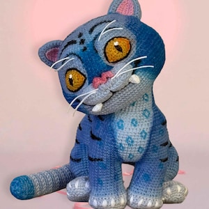 May include: A handmade blue crochet cat with yellow eyes, white whiskers, and a pink nose and ears. The cat has black stripes and spots, and is sitting on a pink background. The cat is a stuffed toy.