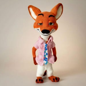 May include: A crocheted fox doll with orange fur, white accents, and a black nose. The fox is wearing a pink shirt, white pants, and a blue tie with a floral pattern. The doll has a smiling expression and is standing upright.