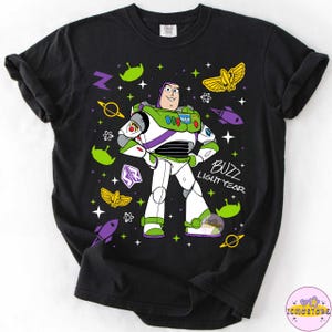 May include: Black t-shirt featuring Buzz Lightyear from Toy Story. The graphic includes Buzz in his spacesuit, surrounded by space-themed elements like rockets, stars, and alien figures. The words "Buzz Lightyear" are printed in white.